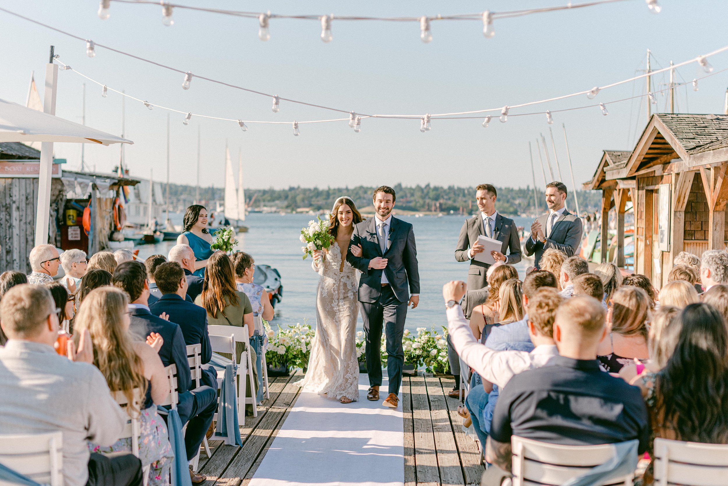 Fine Art Weddings in Seattle, Washington. The Center for Wooden Boats. Bride and Groom at the end of their ceremony, walking down the aisle, while their guests cheer and clap for them. Water rippling in the background with boats here and there.