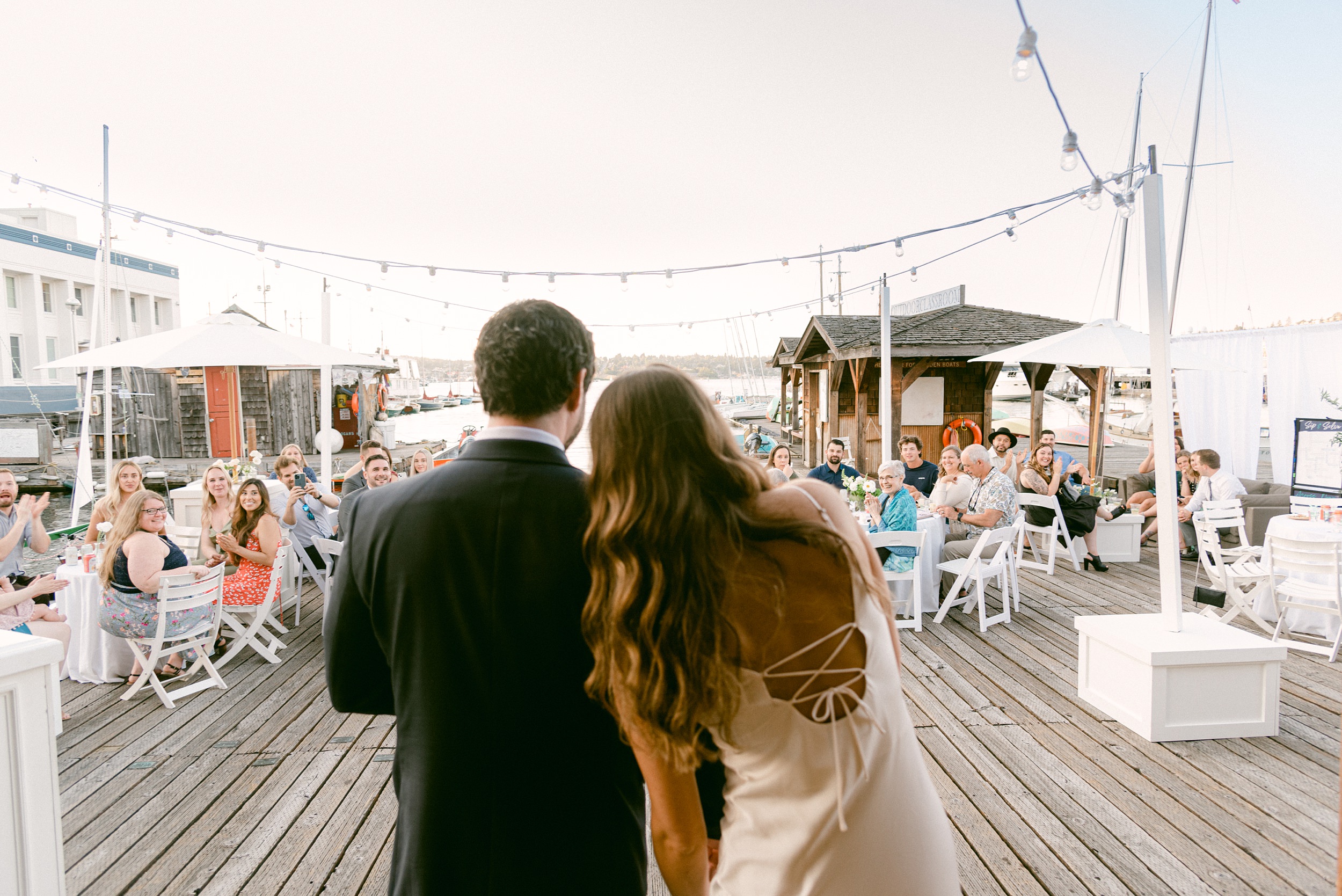 Fine Art Wedding in Seattle Washington. The Center for Wooden Boats. Bride and Groom are announced into reception with loving cheers. They lean on each other in the foreground, while the guests cheer them on in the background. They are facing their guests.