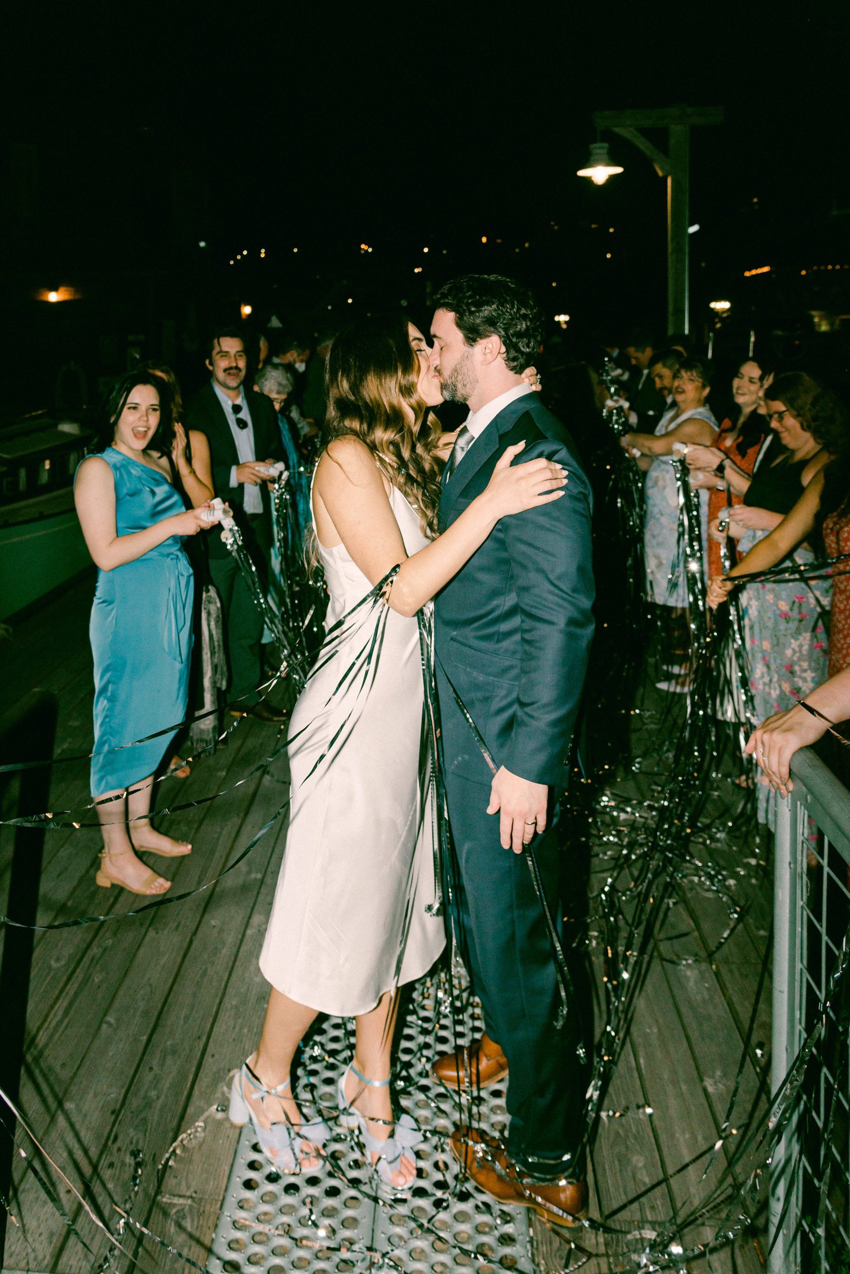 Fine Art Weddings in Seattle Washington. The Center for Wooden Boats, Bride and Groom at their wedding exit, walking through their guests who have confetti streams popping across the image. Bride and groom stop to share a kiss before leaving.